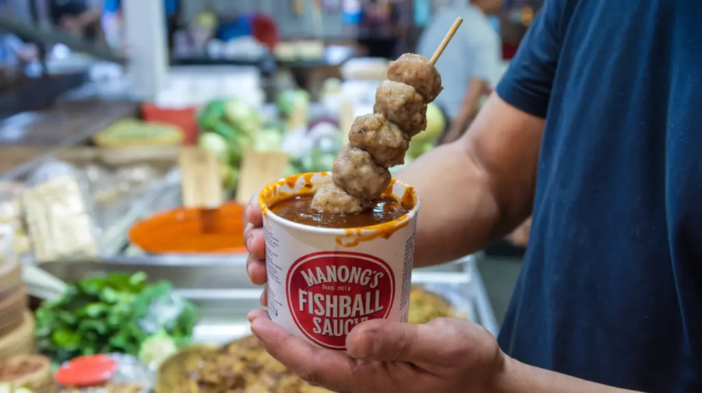 A photo of a man holding a container of Manong's Fishball Sauce. There's a fishball skewer in the sauce. The sauce has a rich, dark color. The background is a food market with various food items.